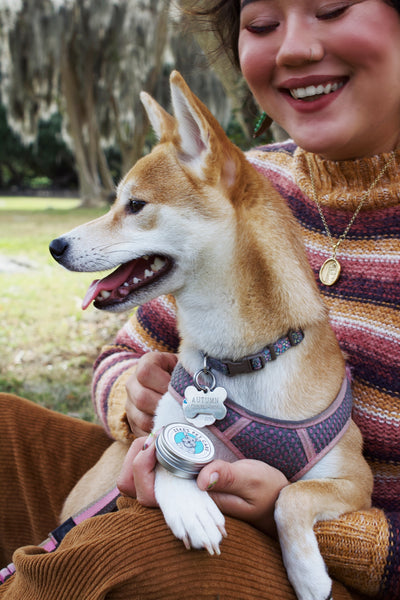 A person with a shiba dog on their lap, person is holding Zekes Soothing Pet Balm. Background depicts mossy trees and grass.
