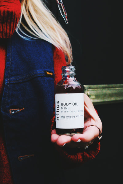 A person with long blonde hair is holding a mint body oil bottle and taking out the eyedropper.