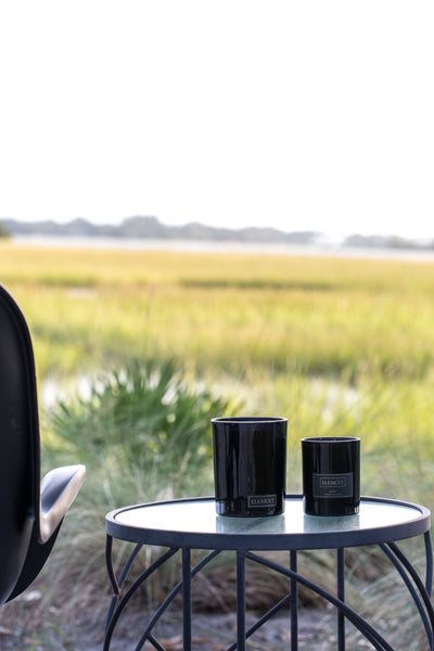 Two different sized candles on a patio table next to a chair. The setting is an outdoor patio/deck area facing a scenic view of the marsh