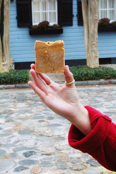 A person's hand holding citrus bar soap. Background depicts cobble stoned streets and window boxes.