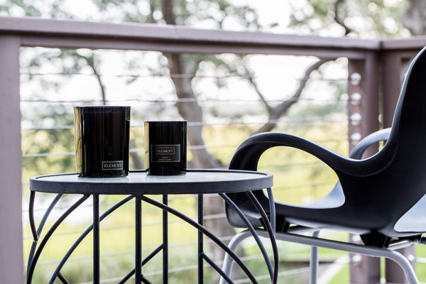 Two different sized candles on a patio table next to a chair. The setting is an outdoor patio/deck area facing a scenic view of trees.